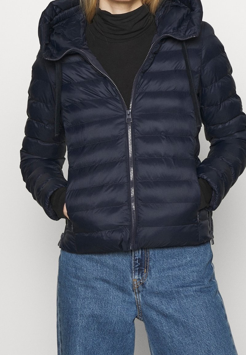 Veste matelassée bleu marine avec fermeture éclair à l'avant, silhouette ajustée, texture matelassée et capuche avec cordon de serrage. Portée sur un col roulé noir.