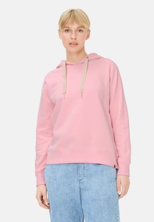 Frau mit kurzen blonden Haaren, die einen hellrosa Hoodie mit beigen Kordeln und hellblaue Jeans trägt, stehend vor einem einfarbigen Hintergrund.