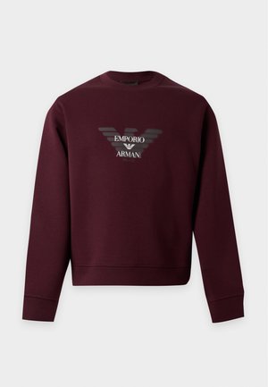 Άνδρας που φοράει μπλουζάκι Emporio Armani σε μαρονοκόκκινο χρώμα, ανοιχτόχρωμα μπλε τζιν και μαύρα παπούτσια, στέκεται με το ένα χέρι στην τσέπη σε λευκό φόντο.