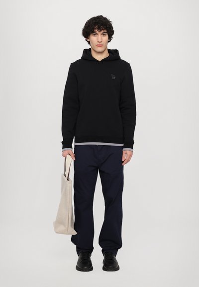Black hoodie with a front pocket and gray hem, paired with navy pants and black shoes. Holding a beige tote bag with brown accents.