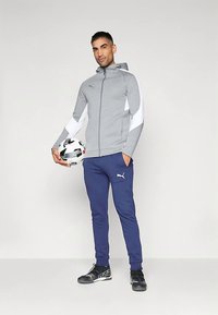 Grå sportsjakke med hvite detaljer, marineblå treningsbukser og svarte sko. Modellen holder en svart-hvitt fotball.