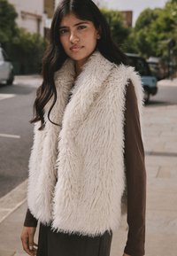 Weiße Kunstpelzweste mit weicher, zotteliger Textur. Verfügt über einen Schalkragen und wird über einem braunen Langarmshirt getragen, vor einem städtischen Hintergrund.