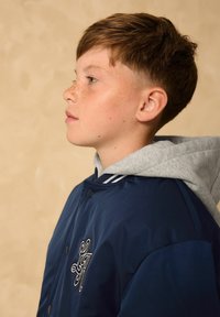 Marineblaue Bomberjacke mit grauem Kapuzenpullover darunter, versehen mit weißen Streifen am Kragen und einem bestickten Design auf der Brust.