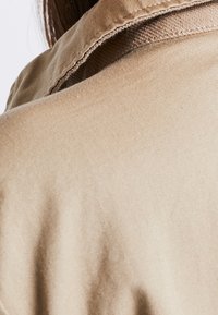 Beige stoffen kraag met stikseldetail, onderdeel van een kledingstuk, toont een gladde textuur en een gestructureerde vorm vanuit de achterkant.