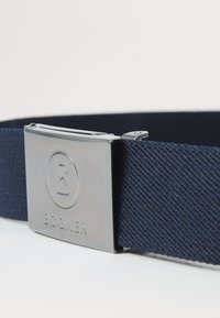 Ceinture en tissu bleu marine avec une boucle en métal argenté brillant, ornée d'un logo circulaire et du nom de la marque "BOGNER" embossé.