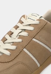 Μπεζ σουέτ αθλητικά παπούτσια με λευκές κορδόνες και ακμές. Διαθέτει υφή στην επιφάνεια, κυματιστές άκρες και διακριτικό λογότυπο. Λεπτή καουτσούκ σόλα.