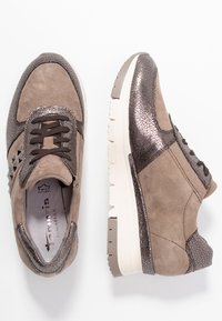 Sko i brunt mocka med metalliska detaljer. Har en strukturerad sidopanel, rund tå och tjock vit sula med ränder.