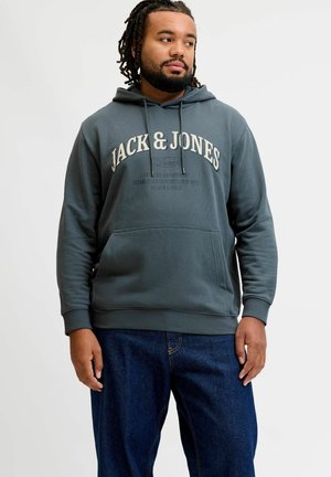 Mężczyzna z dredami, ubrany w ciemnoszarą bluzę z kapturem Jack & Jones oraz niebieskie dżinsy, stojący na jednolitym jasnym tle.