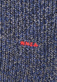 Gebreide stof in marineblauw met witte en zwarte vezels. Heeft een rode geborduurde "NZ"-logo. Gelaagd patroon met een dikke weving.