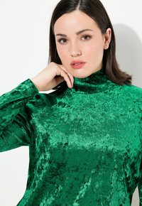 Maglione verde in velluto a collo alto con una finitura lucida e texture; maniche lunghe aderenti; tessuto liscio con un colore ricco e profondo.