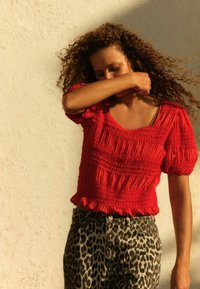 Rød smock-bluse med puffede ærmer og firkantet halsudskæring, parret med leopardmønstrede jeans. Teksturdetaljerne i stoffet er fremtrædende.