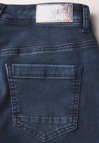 Jean en denim bleu foncé présentant une poche arrière unique, des coutures visibles et une étiquette en métal sur la taille.