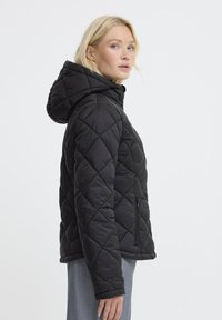 Femme blonde de profil portant une veste à capuche matelassée noire et un pantalon bleu clair, sur fond uni.