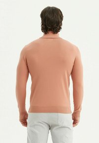 Un pull en maille rose clair avec de longues manches, des poignets et un ourlet côtelés, présentant un design à coupe standard. Porté avec un pantalon de couleur claire.