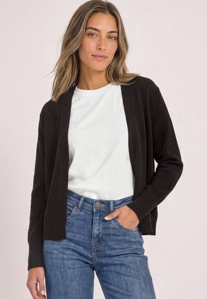 Jonge vrouw met lichtbruin haar draagt een zwarte cardigan, wit overhemd en blauwe spijkerbroek met één hand in de zak tegen een lichte achtergrond.