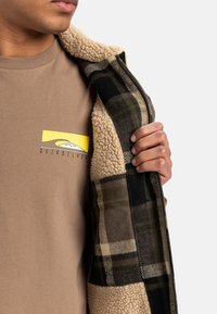 Delvis udsigt til en person iført en tan Quiksilver T-shirt og som holder en sherpa-fodret, grøn og sort ternet jakke åben.