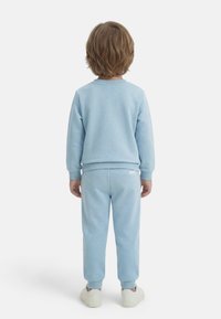 Παιδί με ανοιχτό καστανά μαλλιά, φορώντας ανοιχτό μπλε φούτερ και παντελόνι φόρμας, γυρισμένο από την πλάτη, στέκεται σε λευκό φόντο με λευκά αθλητικά παπούτσια.