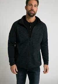 Chaqueta negra con cremallera, fabricada con un tejido texturizado, cuello alto y dos bolsillos laterales. Lleva puesta una camiseta henley negra, combinada con unos vaqueros azules.