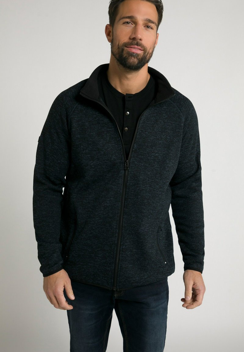 Chaqueta negra con cremallera, fabricada con un tejido texturizado, cuello alto y dos bolsillos laterales. Lleva puesta una camiseta henley negra, combinada con unos vaqueros azules.