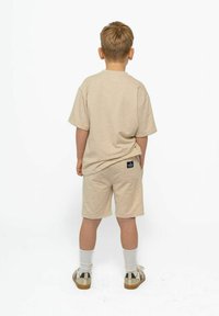 Vaaleanbeige puuvillapaita ja shortsiyhdistelmä, jossa on lyhyet hihat, rento istuvuus, takatasku shortseissa ja pieni etiketti vyötärönauhan kohdalla.