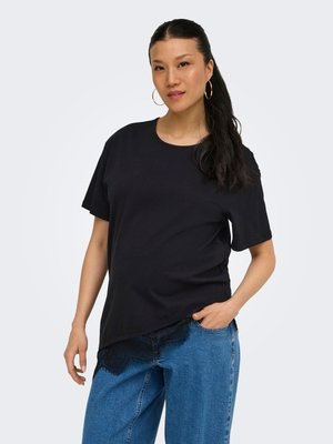 Kvinde med langt mørkt hår iført store hoop-øreringe, en sort kortærmet top med blonderkant og blå jeans, stående med den ene hånd i lommen.