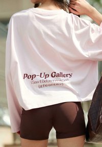 Svetlo roza oversize majica s tekstom na zadnji strani "Pop-Up Gallery I Saw It Before It Was Cool" v temno maroonu, v kombinaciji z rjavimi kratkimi hlačami.