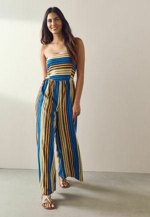 Frau steht in einem trägerlosen, mehrfarbig vertikal gestreiften Jumpsuit mit weitem Bein und trägt Sandalen vor neutralem Hintergrund.