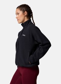 Vrouw met gevlochten haar die een zwarte fleecejas en bordeauxrode leggings draagt, staand in profiel met handen in de jaszakken.