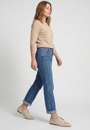 Kvinde gående iført beige sweater, opkørte blå jeans og perforerede beige snøresko mod ensfarvet baggrund.