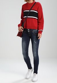 Jersey de punto rojo con rayas en negro y blanco, combinado con unos jeans skinny desgastados en azul oscuro y zapatillas blancas. Acento de bolso cruzado negro.