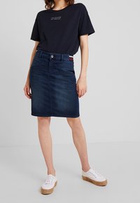 Falda lápiz de denim oscuro con corte recto, que cuenta con un cierre de botón en el frente, agujeros para cinturón y dos bolsillos laterales. Combinada con zapatillas blancas.