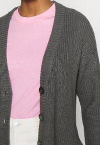 Gray knitted cardigan with ribbed texture, featuring black buttons. Worn over a light pink T-shirt and white pants.