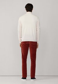Weißer Strickpullover, eng anliegend, kombiniert mit rostbraunen Cordhosen. Das Modell steht vor einem schlichten grauen Hintergrund und trägt marineblaue Sneaker.