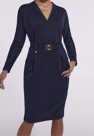 Vrouw die een marineblauwe jurk met lange mouwen draagt, een V-hals en een bijpassende riem met een ronde gouden gesp.
