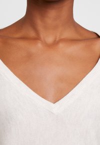 Ljust beige v-ringad tröja med ett texturerat stickmönster och ribbad kant, med en avslappnad passform kring halsringningen och axeln.