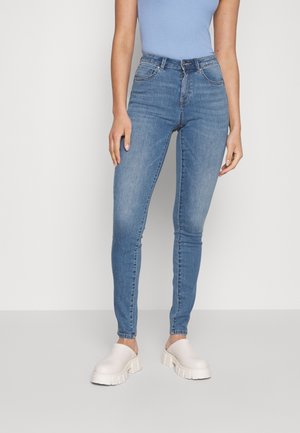 Femme portant un top sans manches bleu clair, un jean skinny bleu taille haute et des chaussures blanches épaisses à enfiler, debout devant un fond uni.