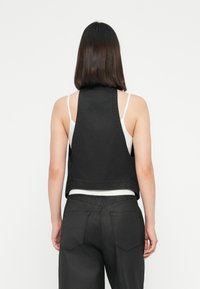Mulher com cabelo escuro na altura dos ombros, usando um colete preto sem mangas sobre uma blusa branca e calças pretas, de costas contra um fundo branco.