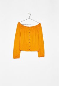 Blouse orange à manches longues et épaules dénudées avec boutons sur le devant, accrochée à un cintre en métal contre un mur blanc.