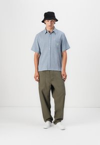 Csíkos, kék rövid ujjú ing, mellzsebbel, olajzöld laza nadrággal és fekete vödörkalappal. A fehér sportcipők teszik teljessé az öltözéket.