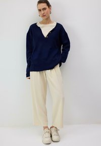 Tmavomodrý pletený sveter s výstrihom do V a tromi hnedými gombíkmi, v kombinácii s krémovými širokými nohavicami a bielymi teniskami.