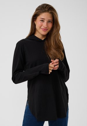 Giovane donna con lunghi capelli castani che indossa una maglia nera a maniche lunghe e jeans blu, sorridente con le mani incrociate davanti.