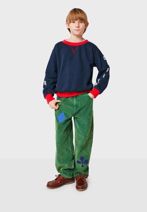 Navyblaue Sweatshirt mit roten Akzenten, kombiniert mit grünen Kordhosen, die blaue und dunkelblaue Patches aufweisen. Braune Schuhe runden das Outfit ab.