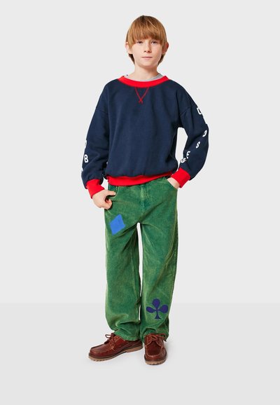 Mörkblå sweatshirt med röda detaljer, kombinerad med gröna kordbyxor som har blå och mörkblå lappar. Bruna skor fulländar looken.