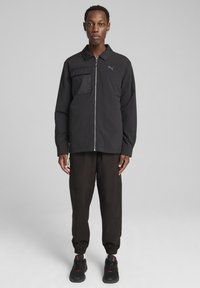 Sort zip-up jakke med en lomme på venstre bryst, parret med løstsiddende sorte bukser og sorte sneakers. Letvægtsstof, minimalistisk design.