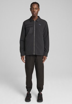 Sort zip-up jakke med en lomme på venstre bryst, parret med løstsiddende sorte bukser og sorte sneakers. Letvægtsstof, minimalistisk design.