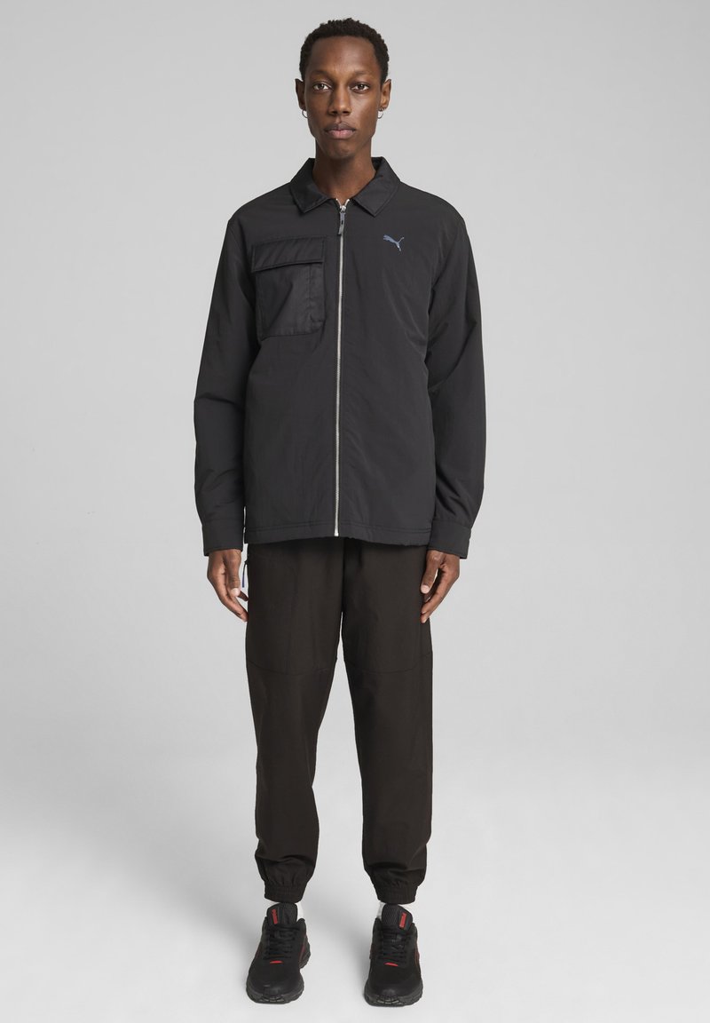 Sort zip-up jakke med en lomme på venstre bryst, parret med løstsiddende sorte bukser og sorte sneakers. Letvægtsstof, minimalistisk design.