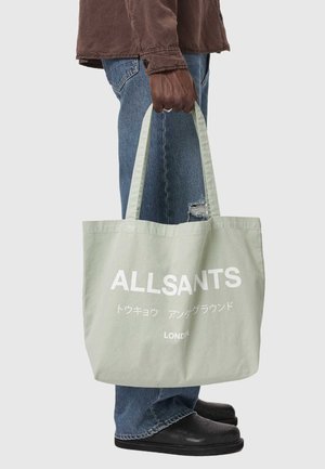 Person hält eine blassgrüne AllSaints-Tasche mit japanischem und englischem Text, trägt ein braunes Hemd, zerrissene blaue Jeans und schwarze Schuhe.