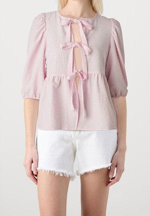 Vrouw draagt een lichtroze blouse met drie strikken aan de voorkant en opgeblazen mouwen, gecombineerd met witte shorts met rafelige zoom.