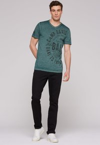 Camiseta verde a rayas con cuello en V, que presenta texto gráfico en negro. Combinada con pantalones negros y zapatillas blancas. Diseño sencillo, estilo casual.