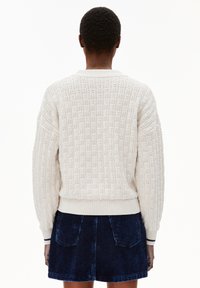 Crème trui met een gestructureerd patroon, geribbelde kraag en manchetten, en een aansluitende tailleband, gecombineerd met een blauwe corduroy rok, gezien vanuit de achterkant.